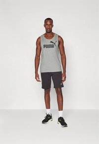 Grå tanktopp med svart "PUMA"-logo, kombinert med svarte shorts. Sorte treningssko med grønne detaljer, står på en ensfarget bakgrunn.