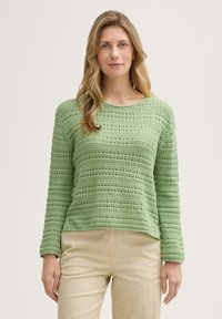 Grüner, locker gestrickter Pullover mit Rundhalsausschnitt, der über strukturierte Muster und lange Ärmel verfügt. Kombiniert mit hellen Hosen.