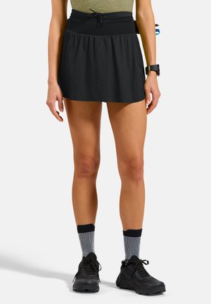 Vrouw die een zwarte sportrok draagt met een verstelbare tailleband, grijze sokken, zwarte hardloopschoenen en een polshorloge, staand tegen een effen achtergrond.