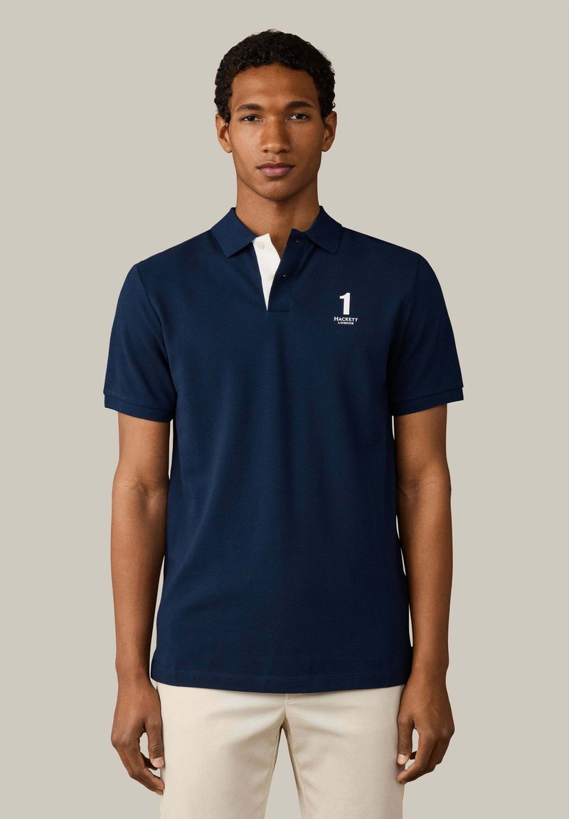Joven con una camisa polo azul marino con tapeta blanca en el cuello y pantalones beige, de pie frente a un fondo gris liso.