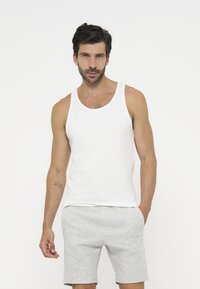 Homme aux cheveux foncés et à la barbe, portant un débardeur blanc et un short gris clair sur un fond uni.