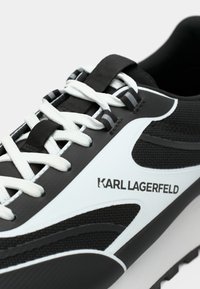 Crno-bijela sportska cipela s teksturiranim gornjim dijelom, kontrastnim naglascima i ravnim bijelim vezicama. Na boku je vidljiv logo.