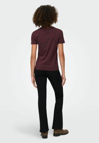 Osoba z kręconymi włosami stoi tyłem, ubrana w bordową koszulę z krótkim rękawem, czarne spodnie i brązowe buty na białym tle.