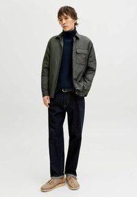 Dunkelgrüne gesteppte Jacke mit einer Brusttasche, getragen über einem dunklen Rollkragenpullover. Kombiniert mit dunklen Jeans und beigen Schuhen. Einfacher, lässiger Look.