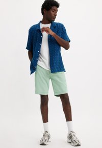 Uomo in camicia a maniche corte blu navy con motivo, T-shirt bianca, pantaloni corti verde menta, calze bianche e scarpe da ginnastica grigie mentre posa su uno sfondo semplice.