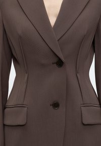 Gros plan d'un blazer marron sur mesure avec deux boutons noirs et des poches à rabat, montrant des coutures structurées et un col à revers.