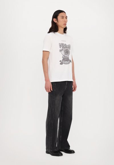 Hvid grafisk t-shirt med en iøjnefaldende trykt design på forsiden, parret med løstsiddende sorte jeans med vertikale sømdetaljer og sorte sko.