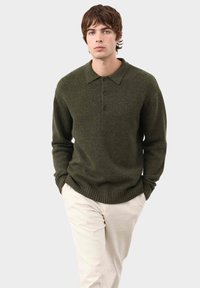Oliwkowy dzianinowy sweter z kołnierzykiem, czterema guzikami i ściągaczami przy hemie i mankietach. Noszony z jasnymi spodniami.