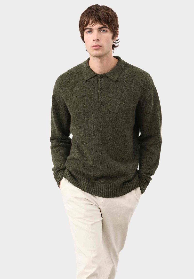 Oliwkowy dzianinowy sweter z kołnierzykiem, czterema guzikami i ściągaczami przy hemie i mankietach. Noszony z jasnymi spodniami.