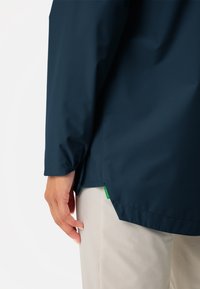 Navy wasserfeste Jacke mit glatter Textur, elastischen Bündchen und seitlicher Schlitze für Belüftung. Enthält ein kleines grünes Etikett am Saum.