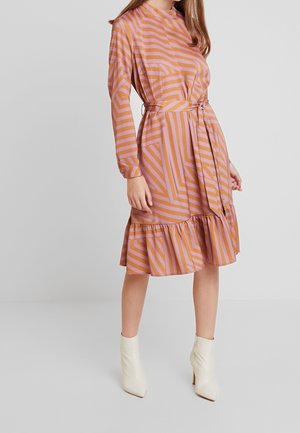 Kobieta ubrana w różowo-pomarańczową sukienkę o geometrycznym wzorze, długości do kolan, z długim rękawem, wiązaną w talii i z falbaną na dole, nosząca białe botki za kostkę.