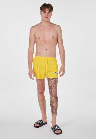 Pantaloni da bagno gialli con vita elastica e coulisse. Indossati con sandali neri con logo bianco. Visibile un notevole tatuaggio su tutta la gamba.