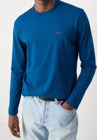 Långärmad blå skjorta med rund halsringning och broderad "BOSS"-logotyp på bröstet, i kombination med ljusa denimjeans och ett svart bälte.