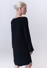 Schwarzes langes Kleid mit gerader Silhouette, weicher Textur und einem dezenten Rundhalsausschnitt auf der Rückseite, kombiniert mit goldenen Ohrringen.