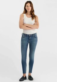 Jean skinny en denim bleu moyen, avec poches avant et fermeture par bouton. Porté avec un débardeur blanc et des mocassins noirs.