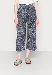 Calças culotte azul-marinho com padrão geométrico branco, cintura alta elástica e cordão, combinadas com sandálias cinzas com tiras ajustáveis.