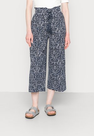 Calças culotte azul-marinho com padrão geométrico branco, cintura alta elástica e cordão, combinadas com sandálias cinzas com tiras ajustáveis.