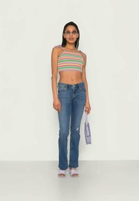 Mulher vestindo um top cropped com riscas arco-íris, jeans azuis, sandálias roxas de salto, óculos de sol e segurando uma pequena bolsa roxa.