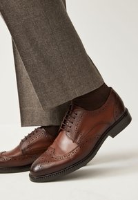 Brown leather dress shoes with wingtip design, decorative brogue detailing, and rounded toe. Worn with brown socks and gray tailored trousers.