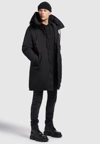 Svart parkas med hög krage, dragkedja och tryckknappar, med ett texturerat inre och sidofickor. Kombinerad med svarta jeans och stövlar.