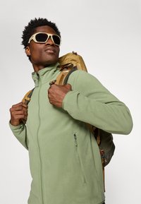Veste en polaire verte avec un col montant et des poches zippées, portée avec un sac à dos beige. De grandes lunettes de soleil blanches complètent le look.