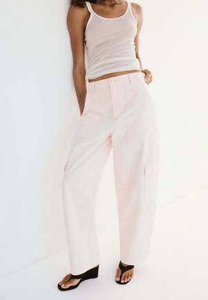 Femme portant un pantalon large rose clair, un débardeur blanc et des sandales compensées noires, debout contre un mur blanc.