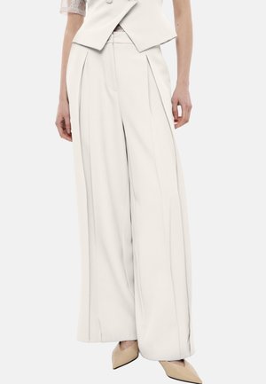 Donna che indossa pantaloni bianchi a vita alta e gamba larga con pieghe anteriori abbinati a décolleté beige con punta affilata.