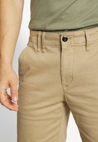 Pantaloni in cotone beige con tasche frontali, passanti per cintura e un bottone nero in vita. Tessuto liscio e taglio dritto.