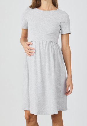 Woman wearing a light gray short-sleeve maternity dress, gently holding her pregnant belly with one hand.