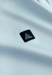 Tissu bleu clair texturé avec une petite étiquette carrée noire arborant un logo triangulaire blanc, placée sur sa surface.