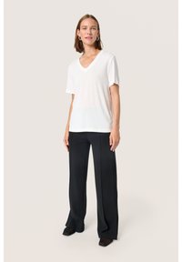Vit v-ringad t-shirt i mjukt material, ihop med svarta vida byxor och svarta klackskor, stående på en neutral bakgrund.