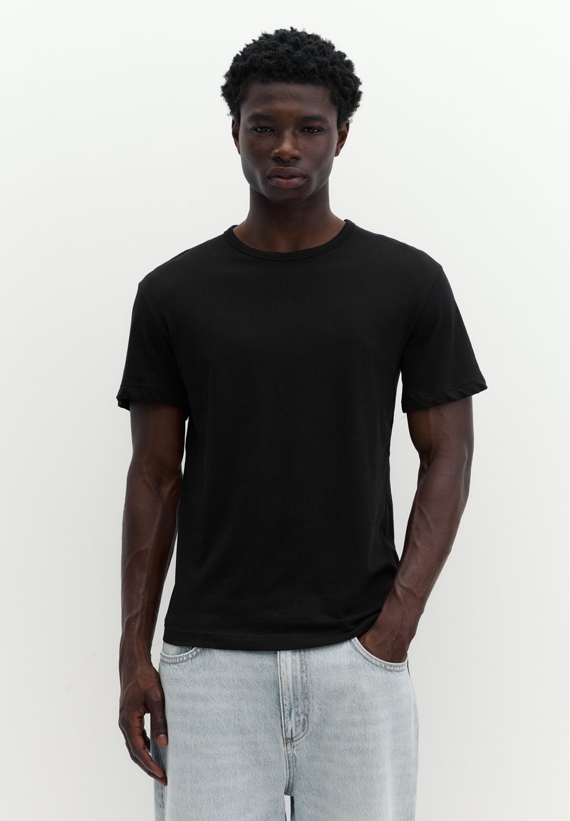 Homme à la peau foncée portant un t-shirt noir uni et un jean bleu clair, debout devant un fond blanc.