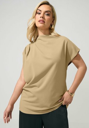 Lyshåret kvinde iført beige kortærmet top og sorte bukser, poserer med den ene hånd på hoften og har gyldt armbånd og øreringe på.