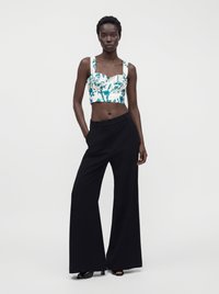 Femme debout, les mains dans les poches, portant un crop top blanc à imprimé floral bleu et un pantalon noir à jambes larges.