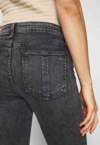 Zbliżenie na osobę noszącą wyblakłe czarne dżinsy z tylną kieszenią oraz beżowy, teksturowany top bez rękawów.