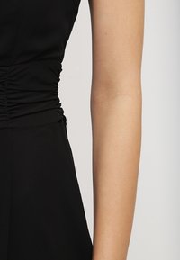 Robe noire avec détails froncés à la taille. Texture lisse et coupe ajustée, mettant en valeur une silhouette élégante. Gros plan sur le bras et la robe.