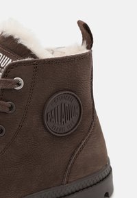 Bottine marron avec un extérieur en cuir texturé, doublure en fausse fourrure, patch logo circulaire et œillets en métal pour les lacets. Semelle en caoutchouc robuste.