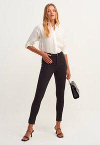 Schwarze, enganliegende Jeans kombiniert mit einem weißen Hemd. Das Model hält eine strukturierte schwarze Handtasche und trägt schwarze Sandalen mit Riemchen.