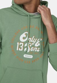 Sudadera verde con un bolsillo tipo canguro en la parte frontal, cordones y un logotipo circular en blanco y naranja que presenta el texto "Only & Sons".
