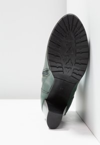 Semelle d'une botte en cuir vert avec une bande de roulement en caoutchouc noir à motifs, posée verticalement contre un mur d'angle blanc.