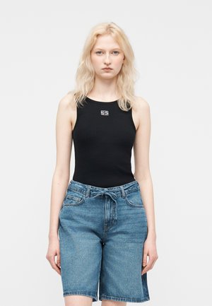 Ung kvinde med blondt hår iført en sort ærmeløs top og højtaljede blå denimshorts foran en ensfarvet lys baggrund.