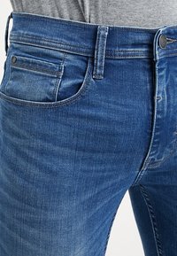 Närbild av blå denimjeans som bärs med en grå topp, visar framfickan, bältesöglor och detaljer med metallknapp.