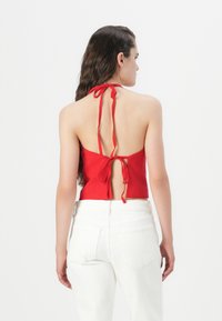 Haut halter rouge avec le dos ouvert, présentant des bretelles à nouer ajustables. Accompagné de jeans blancs taille haute, texture lisse et design ajusté.
