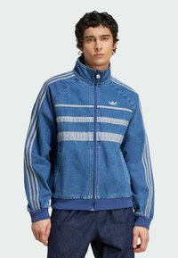 Blå denimjacka med hög krage, framdragkedja och tre horisontella grå ränder. Har ribbade muddar och fåll samt märkets logotyp på bröstet.