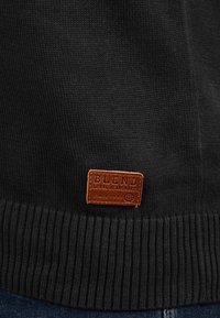 Schwarzer Strickpullover mit geripptem Saum; verfügt über ein rechteckiges braunes Lederpatch mit der Aufschrift "BLEND GLOBAL JEANSMAKER" am unteren Ende.
