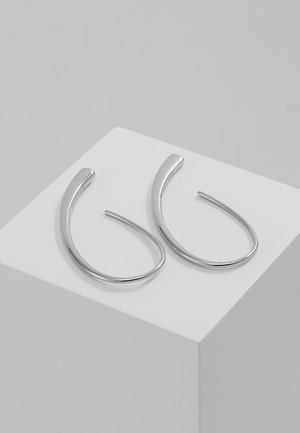 Paire de boucles d'oreilles anneaux en argent minimalistes et courbes présentées sur un cube blanc sur fond gris.