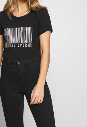 Kobieta w czarnej koszulce z grafiką srebrnego kodu kreskowego i napisem "LIU JO SPORT", w połączeniu z czarnymi, wysokim stanem jeansami.