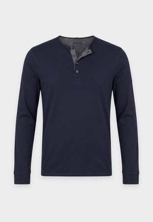 Homme portant une chemise bleu marine à manches longues, un jean foncé avec une ceinture marron et des chaussures en cuir marron, debout devant un fond clair uni.