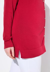 Rote Strickjacke mit geripptem Muster, Knopfverschlüssen und gespaltenem Saum. Die Ärmel sind lang und eng geschnitten, kombiniert mit weißen Hosen.