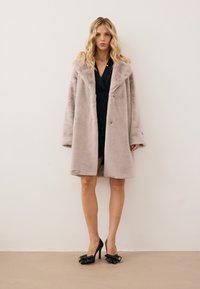 Nep bonten jas in lichtbeige met een grote kraag, dubbele knopen en oversized mouwen. Gedeeltelijk over een zwarte jurk en hoge hakken.